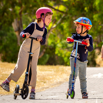 Two small children ride scooters at the park. The child on the left is female, wearing a pink helmet and eggshell coloured overalls on a black, two-wheeled scooter. The child on the right is younger, male and wearing a blue and orange scooter with a navy blue shirt, grey pants and a black scooter with orange handles and green wheels.