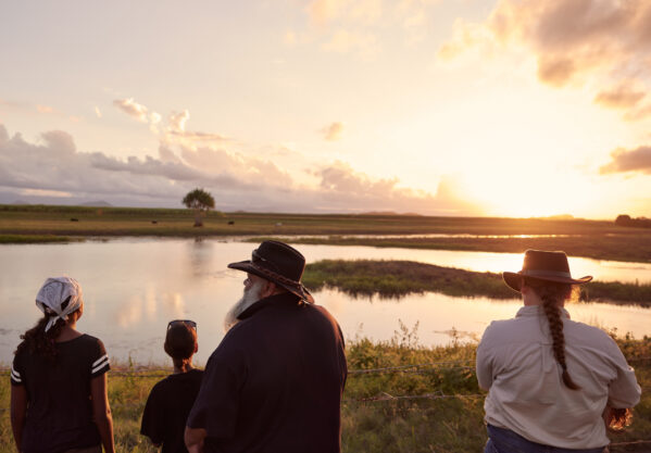 Representatives from Mungalla Station, a Reef Aid program site in Queensland, stand by a wetland overlooking a bright yellow and orange sunset.