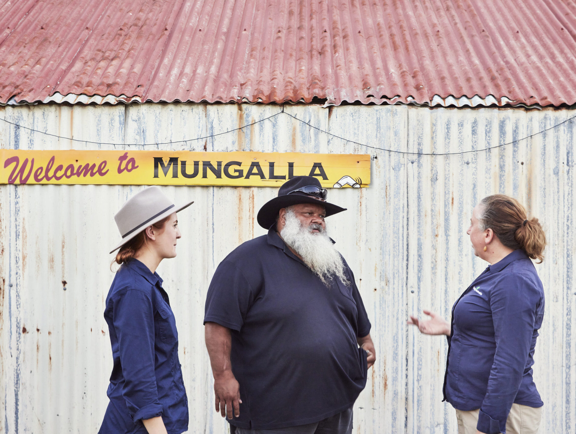 Two female Greening Australia staff members in dark-blue button up shirts speaking to a man in a black cowboy-style hat with a navy blue polo shirt outside Mungalla Station.