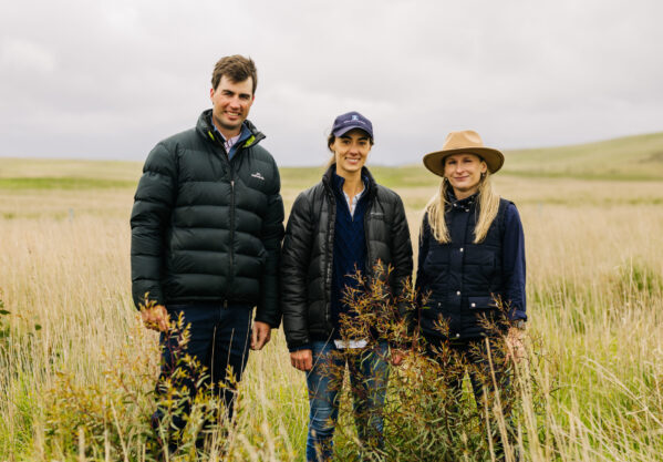 Three people stand in a field of green, slightly dry grass that is knee-height. There is a man on the left with brown, short hair and a black puffer jacket, a woman in the middle with a dark blue cap, black puffer jacket and jeans, and a woman on the right with a tan, wide-brimmed hat,long, blonde hair and a navy blue button-up shirt. All three are smiling.