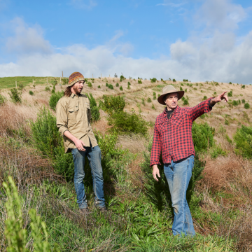 A Greening Australia staff member in a multi-coloured beanie, tan shirt and jeans stands next to a landholder in a red and blue flannel shirt and jeans. The landholder is also wearing a tan wide-brimmed hat, pointing off into the distance.