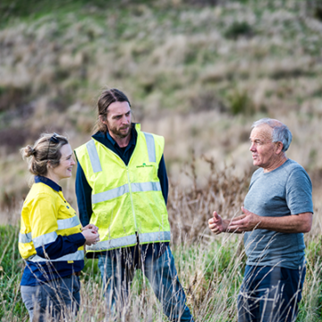 Two Greening Australia staff members - a male and female in high-vis vests and jeans, speak to a landholder on site in Victoria (he is wearing a pale blue t-shirt and dark blue jeans).