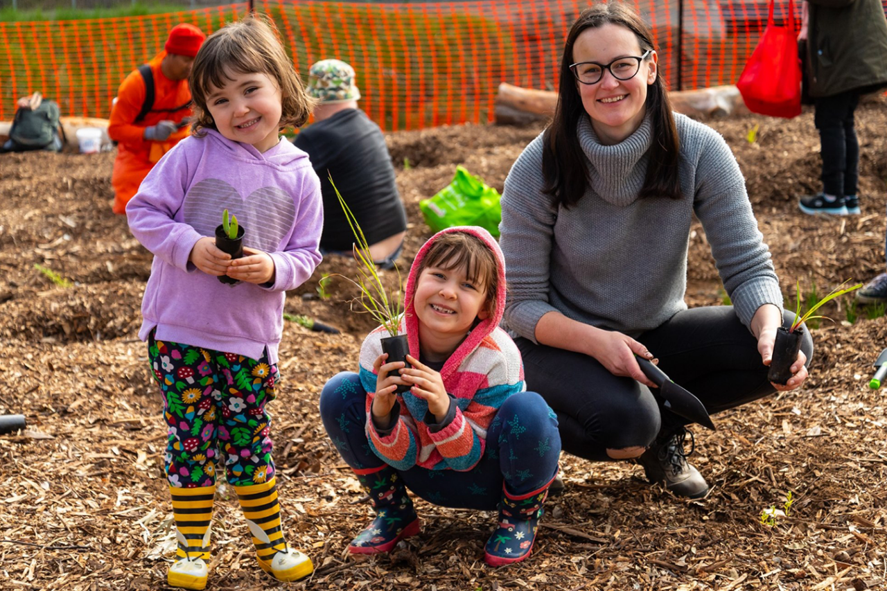 Two small children, both female, with pink hoodies and gum boots, stand holding seedlings next to their mother, who is crouching down beside them. She has long brown hair and is wearing a grey turtle neck. All three are either smiling or mid-laugh.