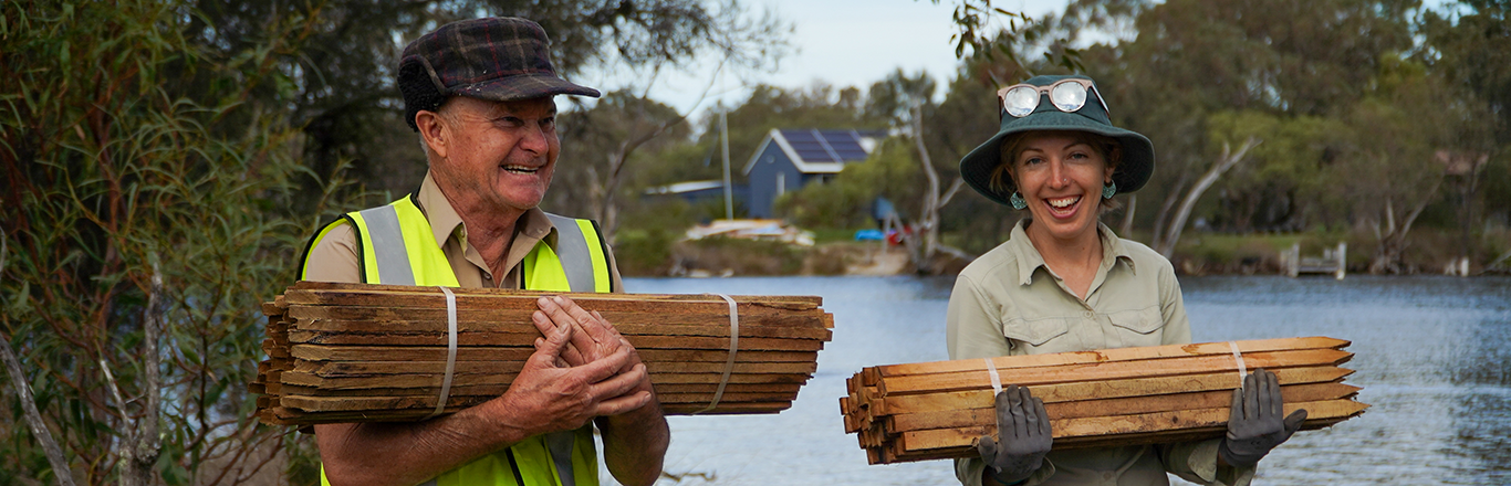 Greening Australia staff member with a green wide-brimmed hat and glasses and volunteer carrying stacks of wood at a planting on Jeegarnyeejip Island. Credit Jessie Collins.