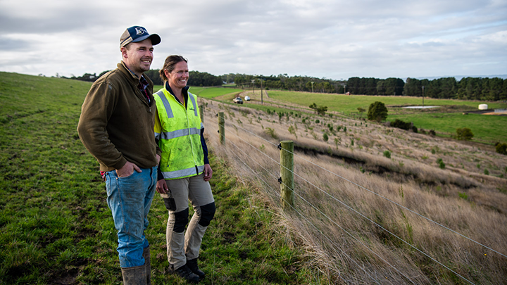 A male landholder in a brown jacket and light blue denim jeans with a cap stands next to a female Australian staff member in a high-vis shirt, both overlooking the landscape of the landholder's property.