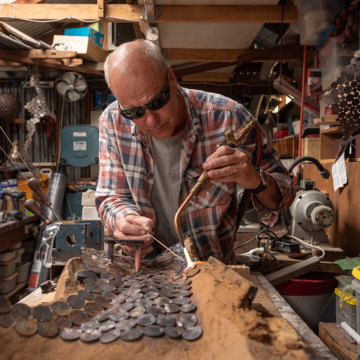 Artist Michael Moerkerk welding two recycled coins together in his workshop. Michael is wearing a flannel shirt and black safety goggles, surrounded by various tools placed on metal racks across wooden walls. Image by Christopher Chan.