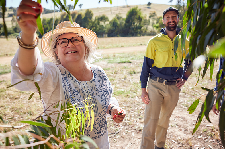 A female landholder in a white shirt and tan, wide-brimmed hat reaches for a healthy, green leaf. Accompanied by a male Greening Australia staff member, laughing, with a yellow, high-vis shirt and tan pants.