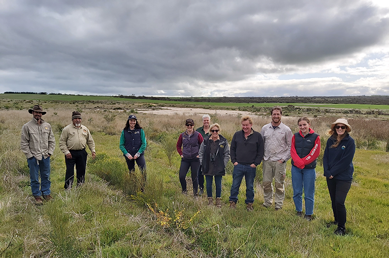 A group of project participants stand in a large group, smiling at the camera, amongst a luscious green filled on a cloudy day.