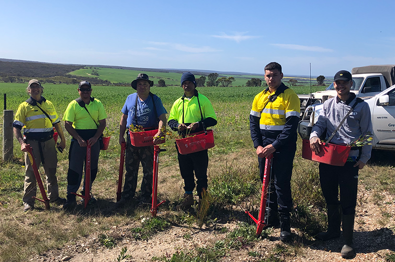 Nowanup Noongar Rangers pose for the camera with their bright red, waist-height seeding machines; wearing fluorescent yellow working shoes, blue pants and brown, steel-capped boots. 
