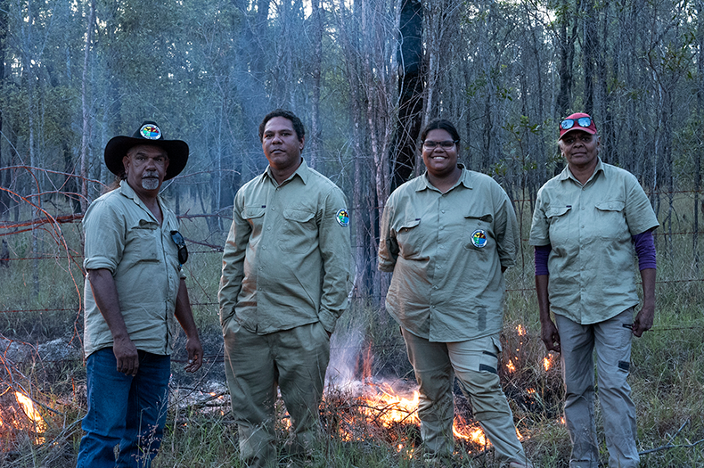 Newly appointed Woorabinda Rangers attend a cultural burning, standing proudly in front of the healthy bush landscape.