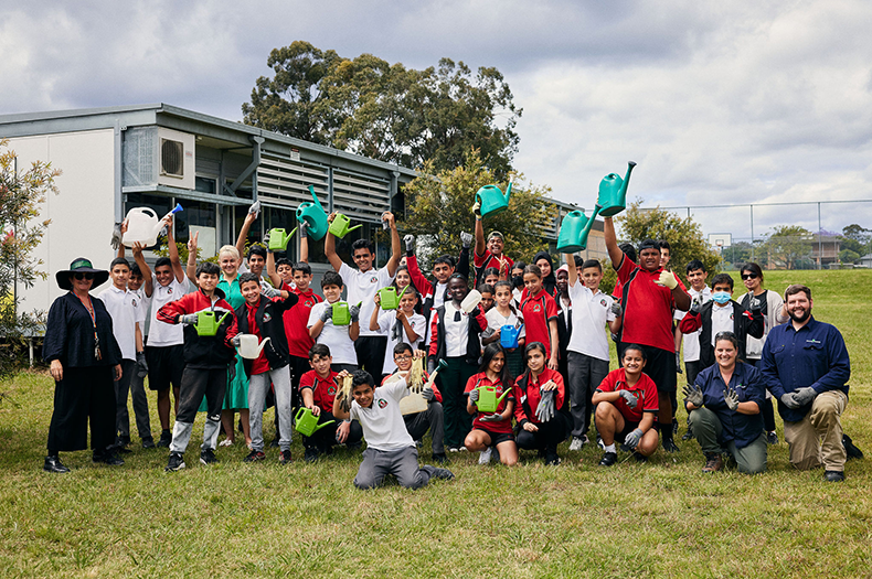A class of students stand in a group on the grass, raising watering cans triumphantly in the air. The class is flanked by their teacher on one side, and two Greening Australia staff on the other.
