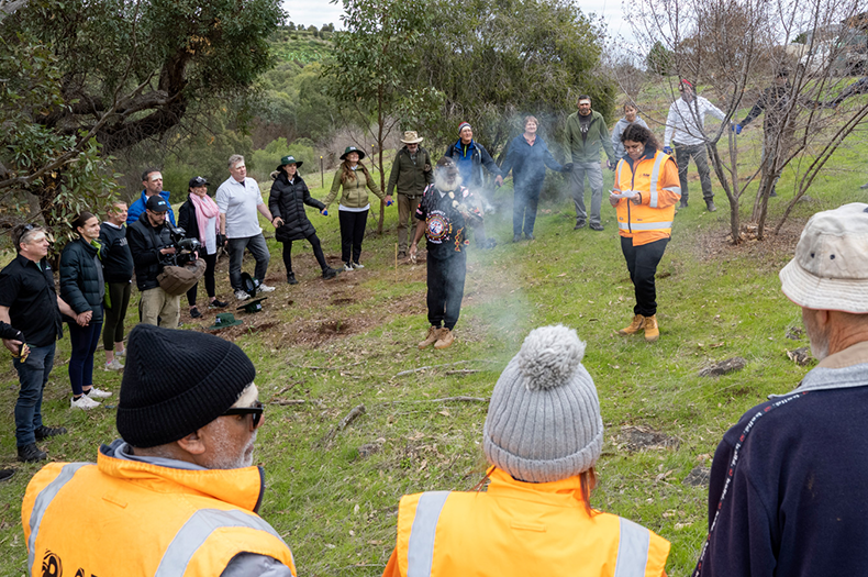 Two Kaurna representatives conduct a smoking ceremony and welcome to Country on a hillside, standing in the middle of a circle of people holding hands..