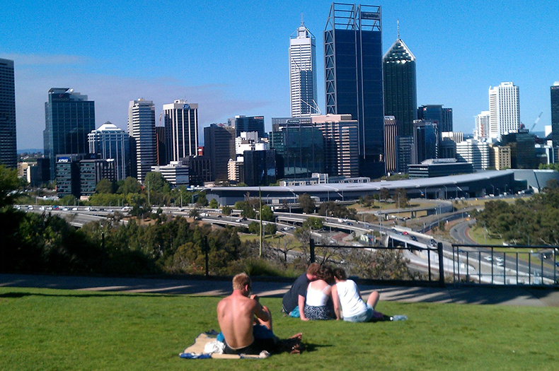 People are sitting relaxing on a public lawn in the foreground, with their backs to the camera, looking towards a city full of skyscrapers and high-rise buildings in the background.