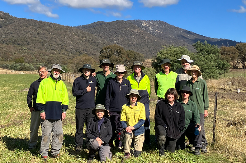 A group of people stand in a paddock, posing for the camera. There are wooded hills in the background and just visible to the right of the group is a replanted area with tree guards visible.