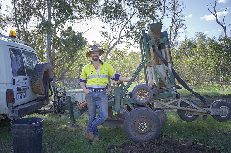 A man stands out in a paddock, smiling at the camera. He is leaning on a tree seeder, hooked up to a ute.