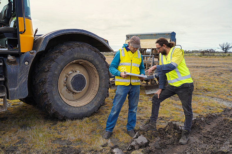 Two people wearing high vis stand out in a paddock, engrossed in discussing a document that one of them holds. Behind them is a large tractor with a seeding machine hitched to the back. 