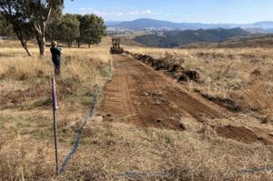 A earthmoving machine is seen in the distance, having scraped the top layer of soil away, leaving a strip of bare earth in a paddock.